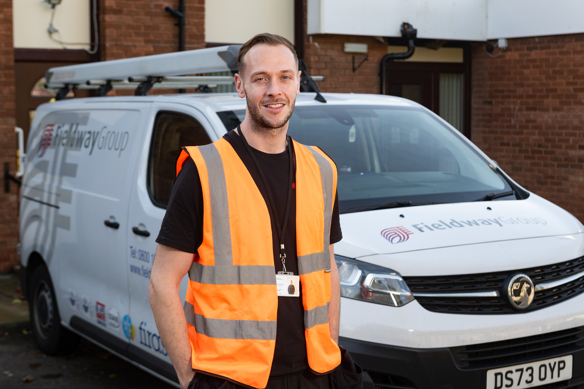 Man with an orange vest and a black tshirt smiling. Behind is a white van with a fieldway logo on it.